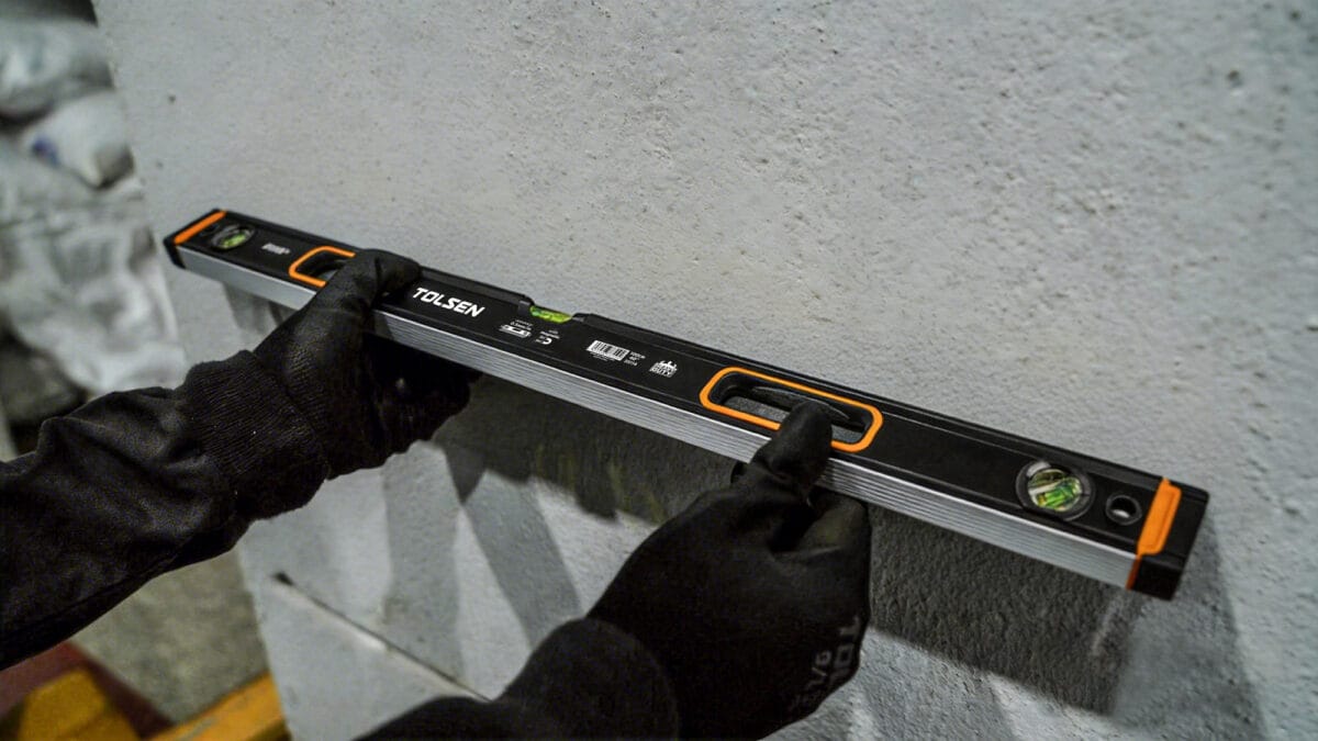 Worker wearing gloves using the TOLSEN magnetic spirit level to check horizontal alignment against a concrete wall.