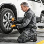 Mechanic wearing Tolsen work jacket and pants kneeling while changing a car tire.