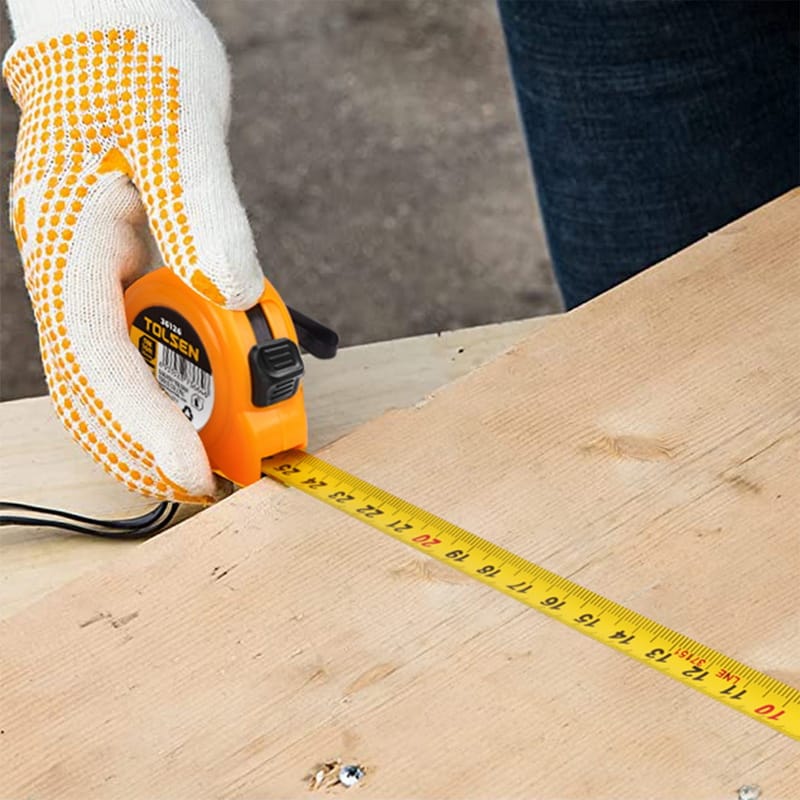 Construction worker wearing gloves measuring a wooden plank using a Tolsen yellow tape measure.