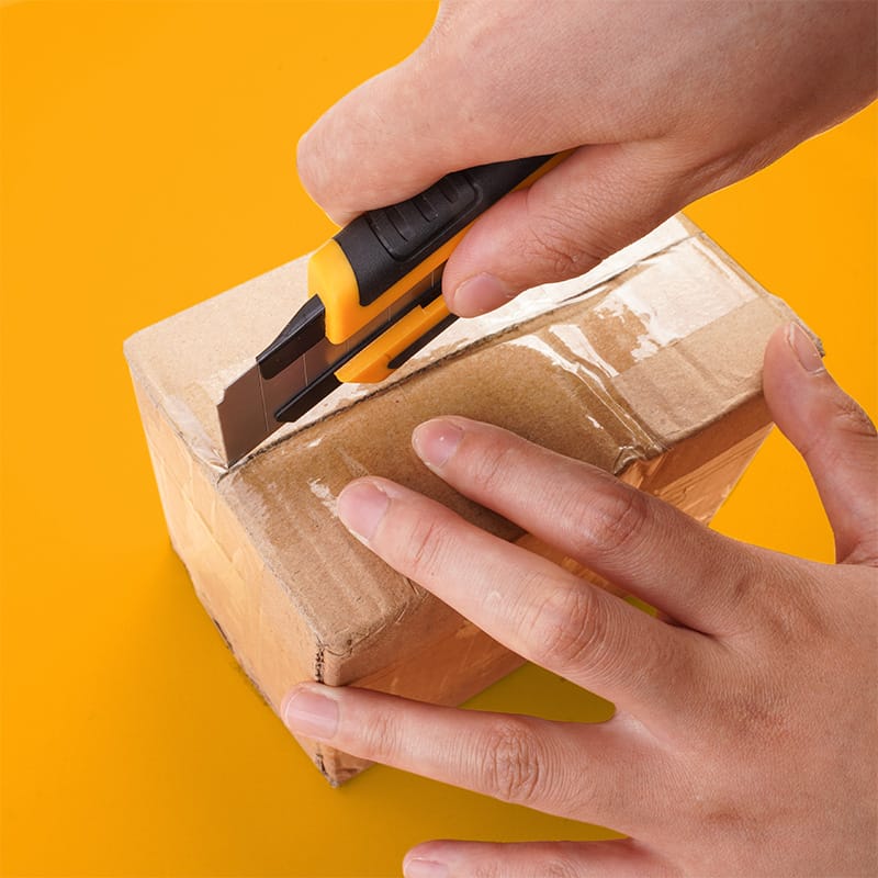 Tolsen utility knife being used as a box cutter to slice through clear packaging tape on a cardboard carton.